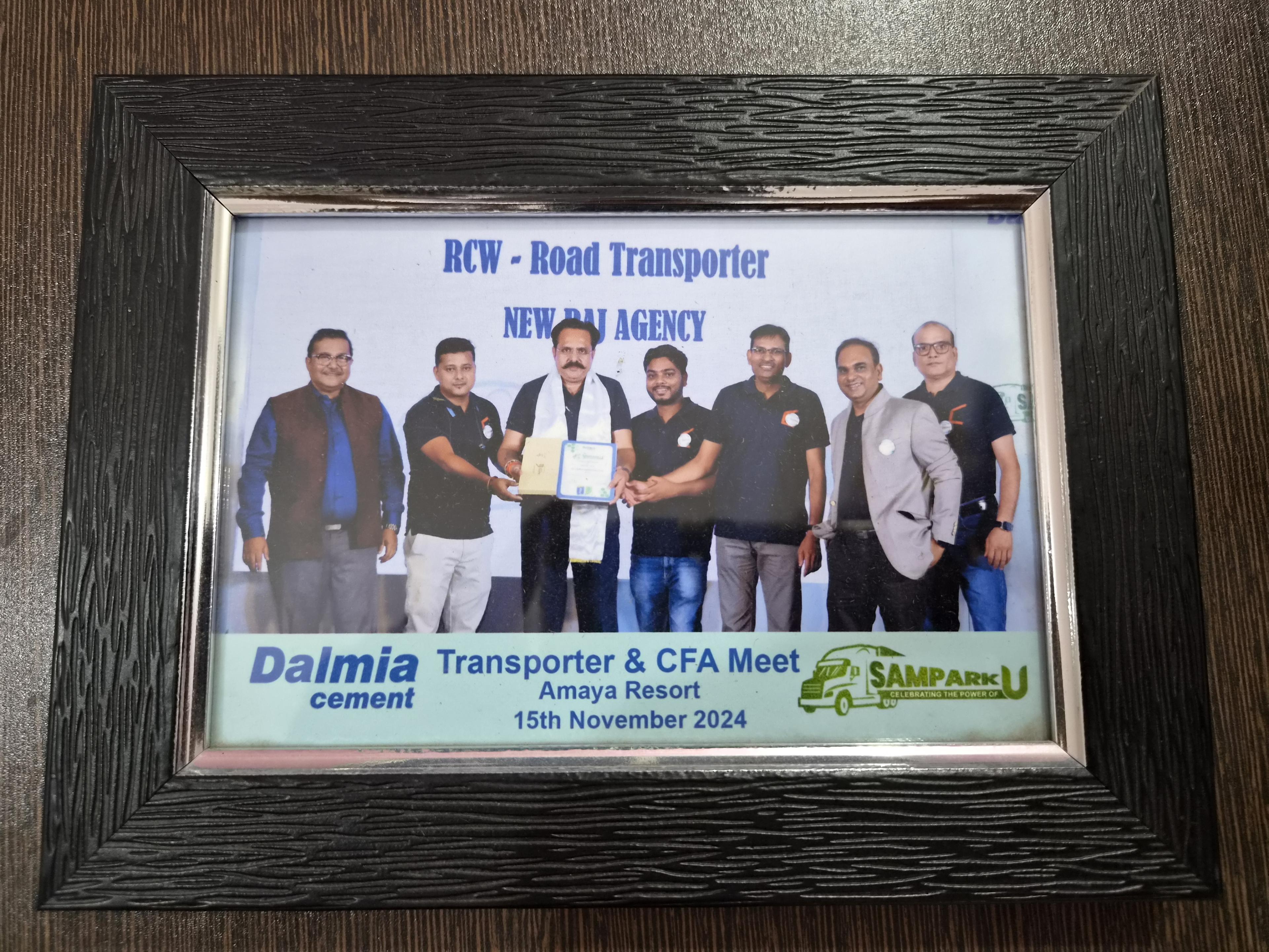 A framed photograph from the Dalmia Cement Transporter and CFA Meet held at Amaya Resort on 15 November 2024, showing attendees gathered for a presentation